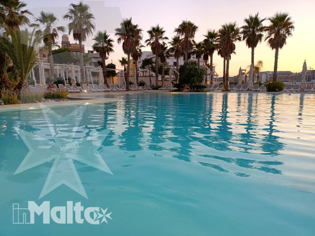 Piscina esterna del resort tra palme e lettini al tramonto Ampia piscina esterna con acqua turchese, palme e lettini nella luce calda del tramonto, ambiente ideale per il tempo libero a Malta.