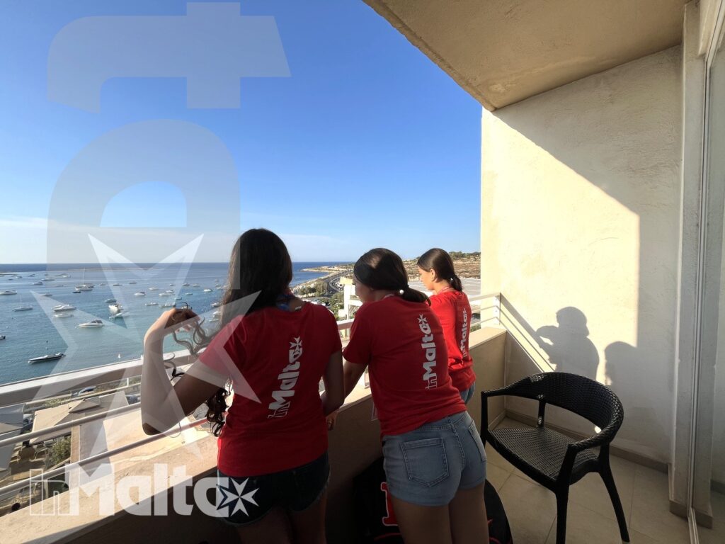 Tre studentesse osservano una baia dal balcone, appoggiate alla ringhiera tra sedie da esterno, barche e costa urbana luminosa.