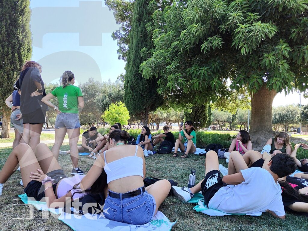 Gruppo internazionale di studenti si rilassa sul prato ombreggiato con abbigliamento estivo, vivendo insieme un'attività all'aperto a Malta.