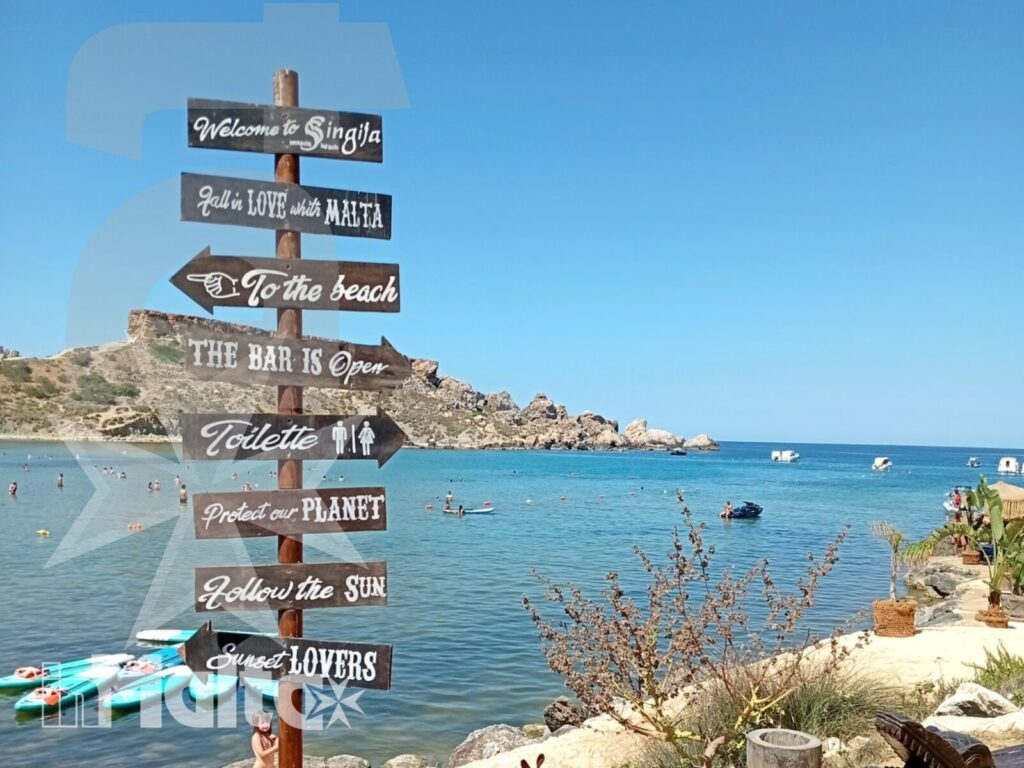 Cartello panoramico e kayak sulla costa maltese Un cartello decorativo indica la baia mediterranea sullo sfondo, dove kayak colorati sostano vicino alla costa rocciosa e all'acqua turchese.