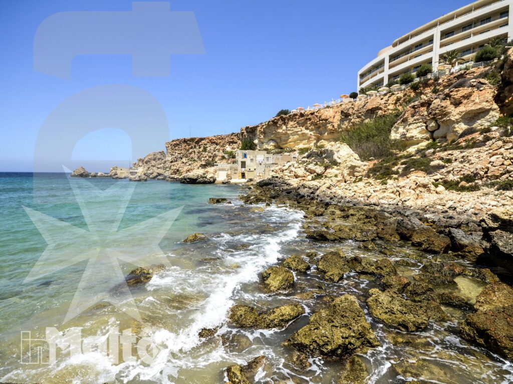 Costa di Golden Bay con struttura sul promontorio Costa rocciosa di Golden Bay con mare turchese, scogli lambiti dalle onde e struttura ricettiva sul promontorio sotto il cielo limpido.