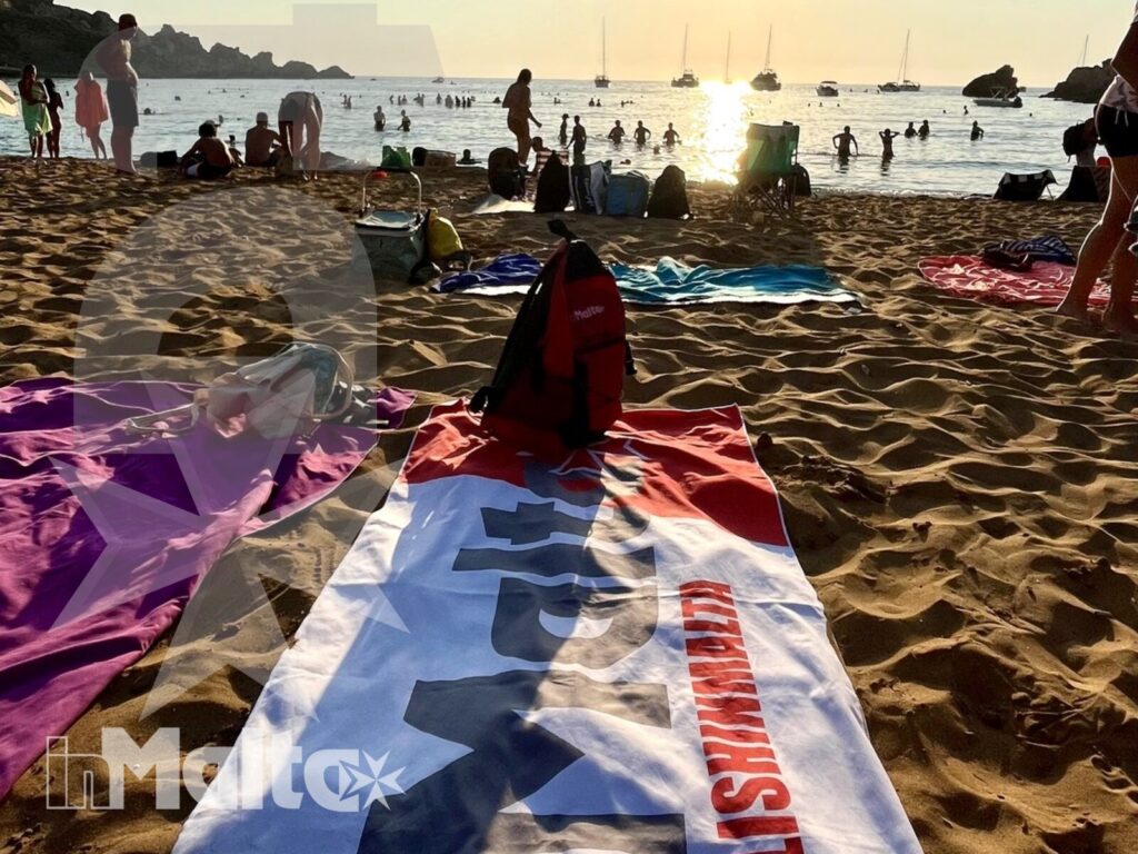 Telo inMalta sulla spiaggia durante il tempo libero degli studenti Telo promozionale sulla sabbia davanti a studenti e bagnanti, con il mare e la scogliera illuminati dal tramonto.