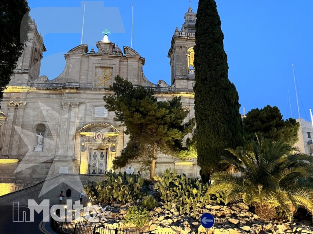 Cattedrale monumentale e giardino mediterraneo inMalta Cattedrale monumentale illuminata accanto a un giardino mediterraneo con cipresso e piante grasse, tappa serale di un itinerario inMalta.