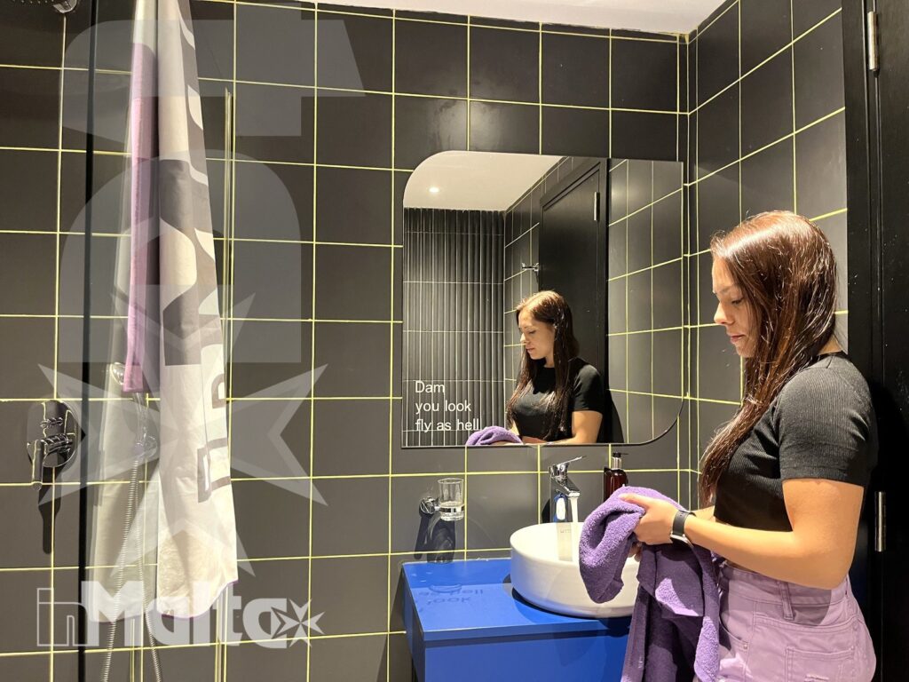 Bagno moderno dell'alloggio con asciugamani e lavabo colorato Donna sistema un asciugamano viola davanti al lavabo blu, in un bagno moderno con piastrelle scure, doccia e specchio ovale.