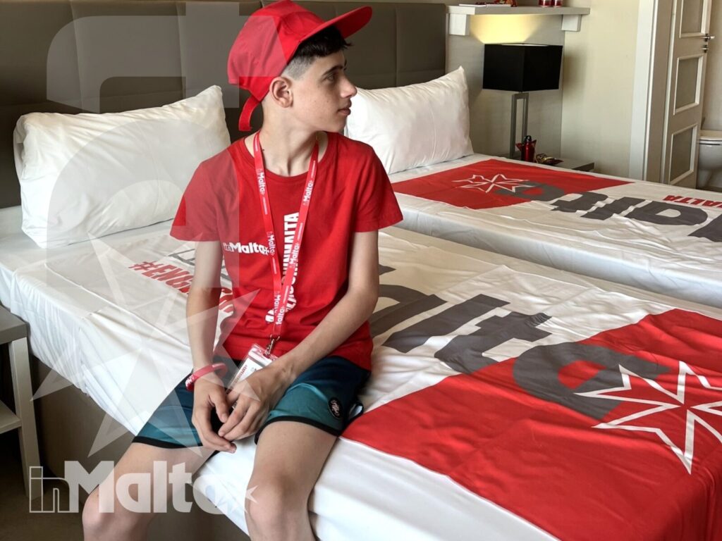 Un giovane partecipante siede sul letto con cappellino e maglietta rossi, mostrando l'esperienza quotidiana in una camera del soggiorno studio inMalta.
