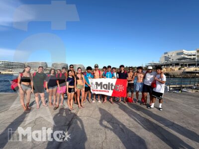 Gruppo internazionale di studenti posa con uno striscione sul lungomare di Malta, davanti al porto e al cielo limpido.