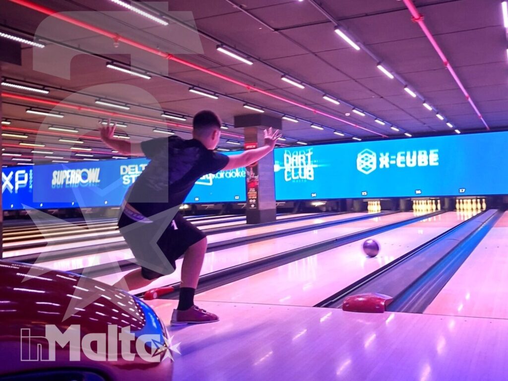 Giovane partecipante lancia la palla lungo una pista illuminata, tra luci colorate e display in un centro bowling moderno.