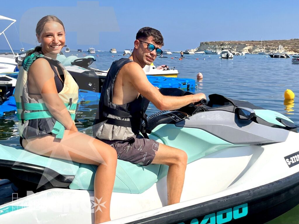 Due studenti sorridenti siedono su una moto d'acqua in rada, con boe, barche ancorate e costa rocciosa alle spalle.