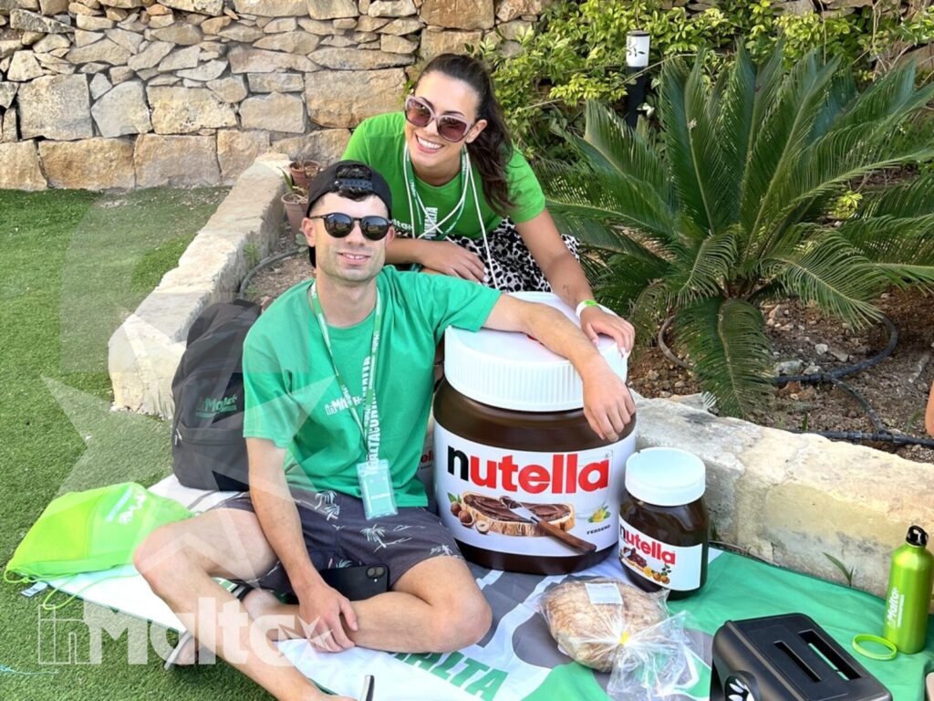 Due studenti sorridenti condividono un picnic sul prato, con crema spalmabile e attrezzatura per attività all'aperto.