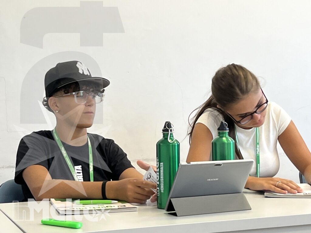 Partecipanti al lavoro collaborativo durante un soggiorno linguistico Due giovani partecipanti collaborano al tavolo usando tablet e quaderni, con badge e borracce coordinate del programma.