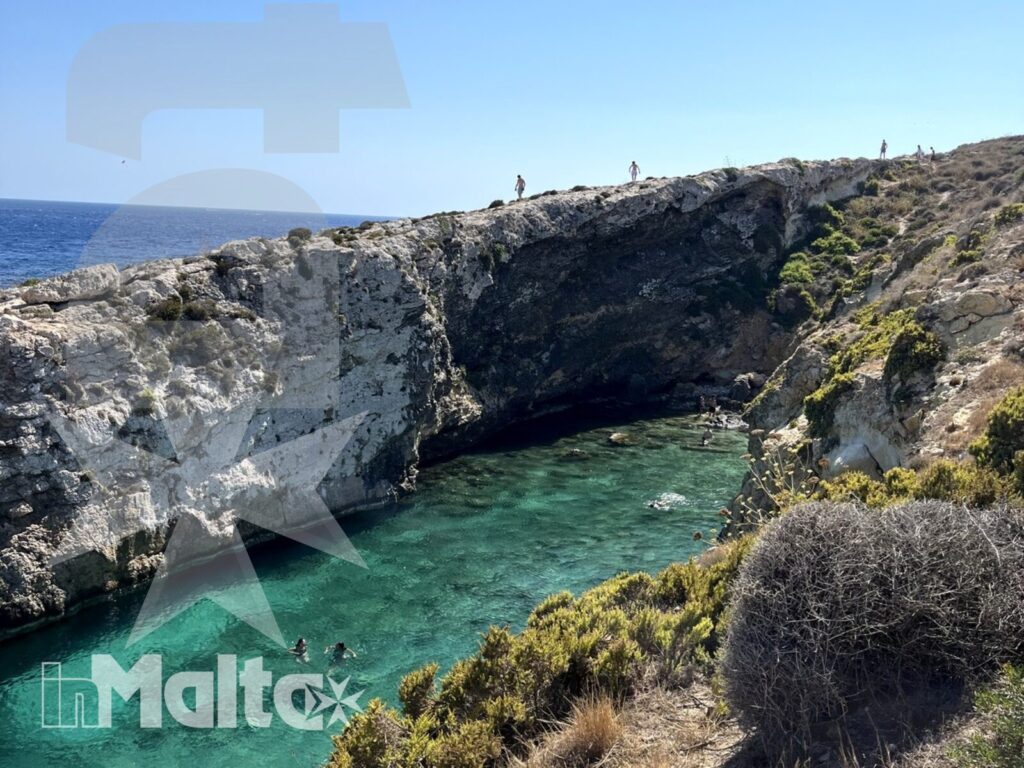 Acque turchesi e scogliere dell'insenatura di Hondoq Bay Veduta panoramica di un'insenatura mediterranea dalle acque turchesi, tra scogliere calcaree, vegetazione costiera e persone in acqua.