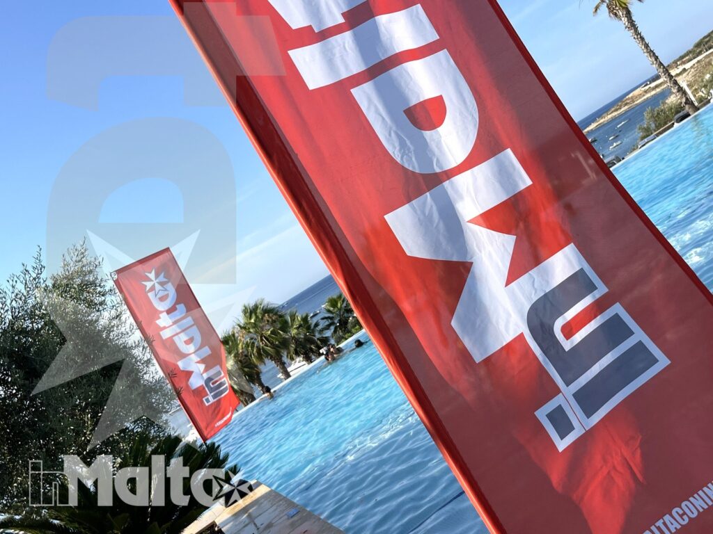 Bandiere rosse accanto alla piscina azzurra del resort, con palme e mare sullo sfondo in una giornata soleggiata.