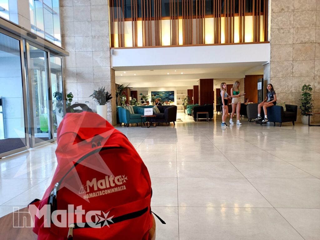 Zaino rosso con logo Malta nell'atrio moderno, mentre alcuni studenti siedono nelle aree comuni illuminate dalle ampie vetrate.