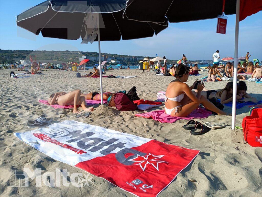 Giornata estiva sulla spiaggia di Mellieha Bay Spiaggia affollata con persone sotto gli ombrelloni, un grande telo promozionale in primo piano e la costa maltese sullo sfondo.