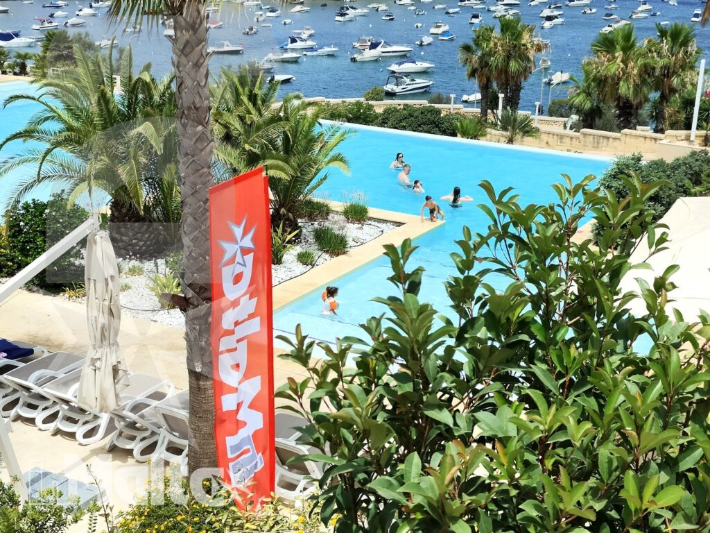 Piscina del resort con vista sul porto di Malta Piscina azzurra del resort con bagnanti, palme, lettini e barche visibili nel porto maltese.