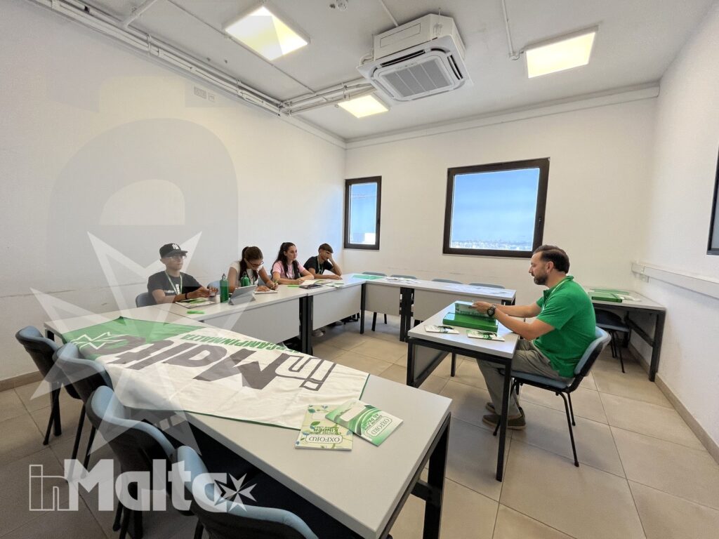 Docente in polo verde conduce una lezione, mentre gli studenti ascoltano ai tavoli disposti a U nell'aula luminosa.