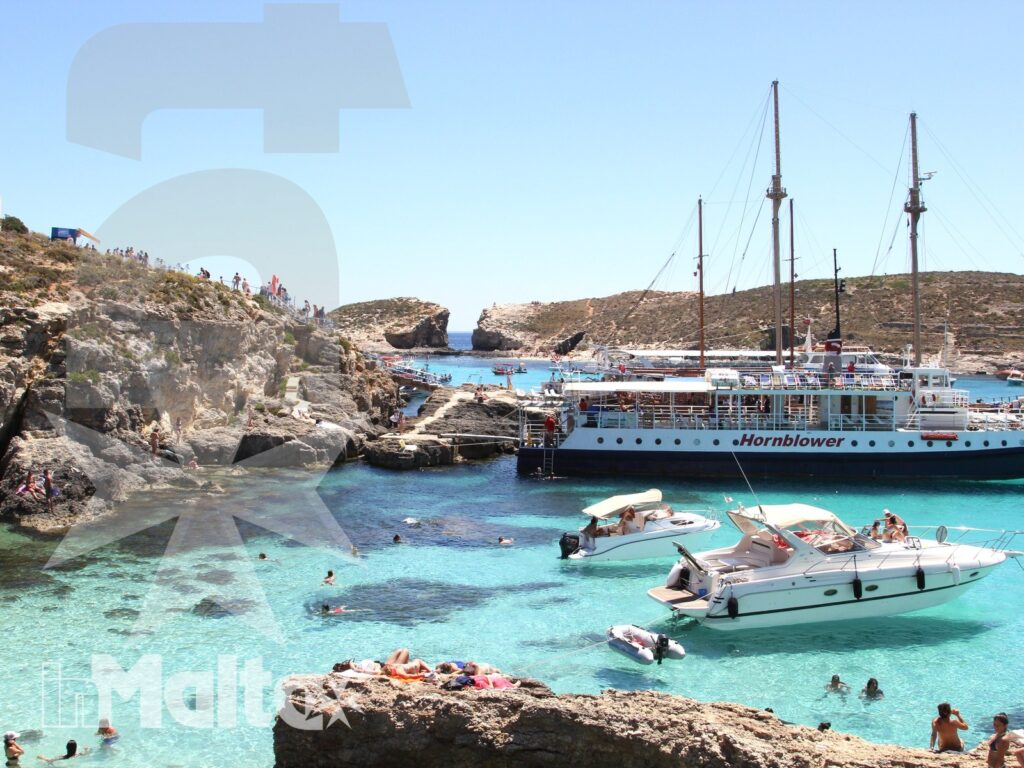 Barche e bagnanti nella Blue Lagoon di Malta Veduta ampia della Blue Lagoon con barche turistiche, nuotatori e mare trasparente tra le rocce mediterranee.