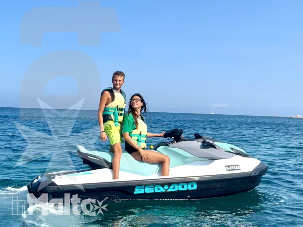 Due giovani con giubbotti salvagente sorridono su una moto d'acqua nel mare aperto, con cielo sereno e costa all'orizzonte.
