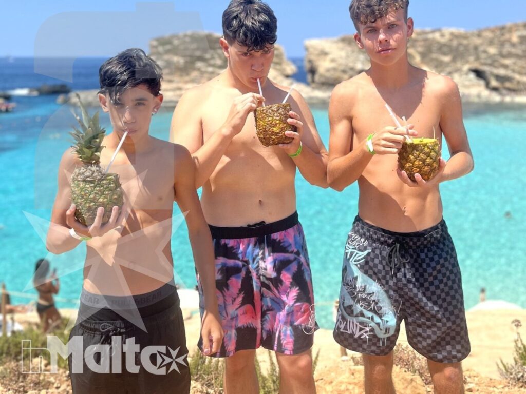 Tre studenti mostrano e bevono da ananas sulla costa maltese, con mare turchese e atmosfera conviviale.