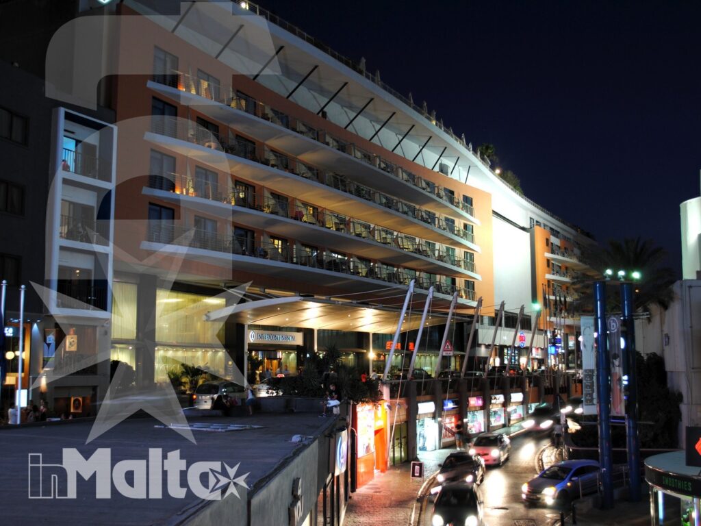 Hotel e strada urbana illuminata nella notte di Paceville Hotel moderno illuminato, con balconi, ingresso coperto e strada trafficata nel vivace contesto serale di Paceville.