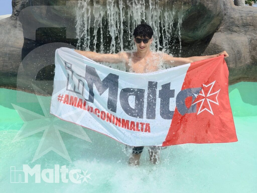 Partecipante in piscina sotto la cascata durante l'attività a Malta Un giovane partecipante immerso in piscina mostra una bandiera promozionale sotto una cascata artificiale, durante una giornata di svago a Malta.