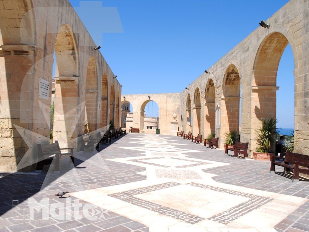 Cortile storico maltese con doppie arcate in pietra, pavimentazione geometrica, panchine e scorcio del mare sullo sfondo.