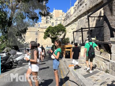 Studenti camminano e conversano sul lungomare di Malta, tra il Mediterraneo, scooter parcheggiati e antiche mura in pietra.