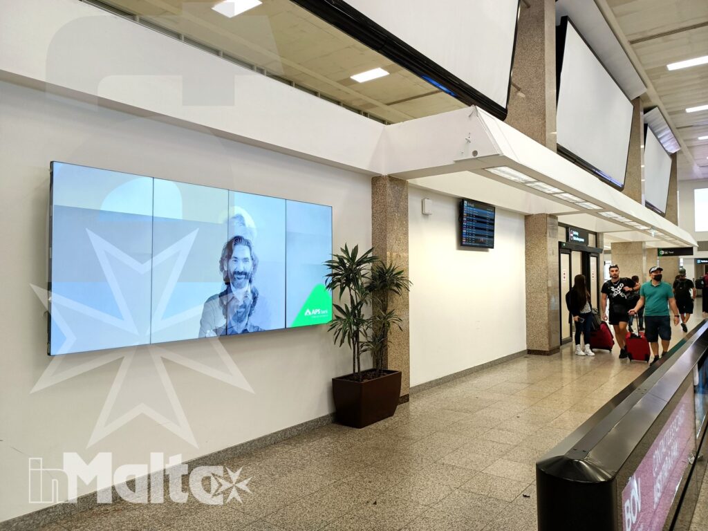 Viaggiatori e bagagli nel corridoio degli arrivi aeroportuali Corridoio moderno dell'aeroporto con viaggiatori, bagagli, nastro trasportatore e schermo digitale promozionale.