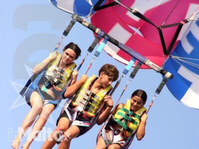 Tre giovani volteggiano durante il parasailing, sostenuti da imbracature e cinghie sotto un grande paracadute blu, rosa e bianco.