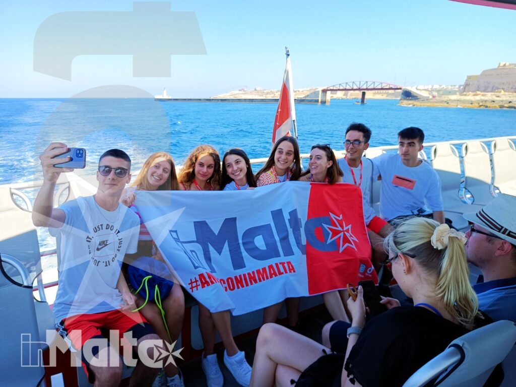 Studenti sorridenti mostrano la bandiera maltese durante una gita in barca, con la costa e un ponte sullo sfondo.