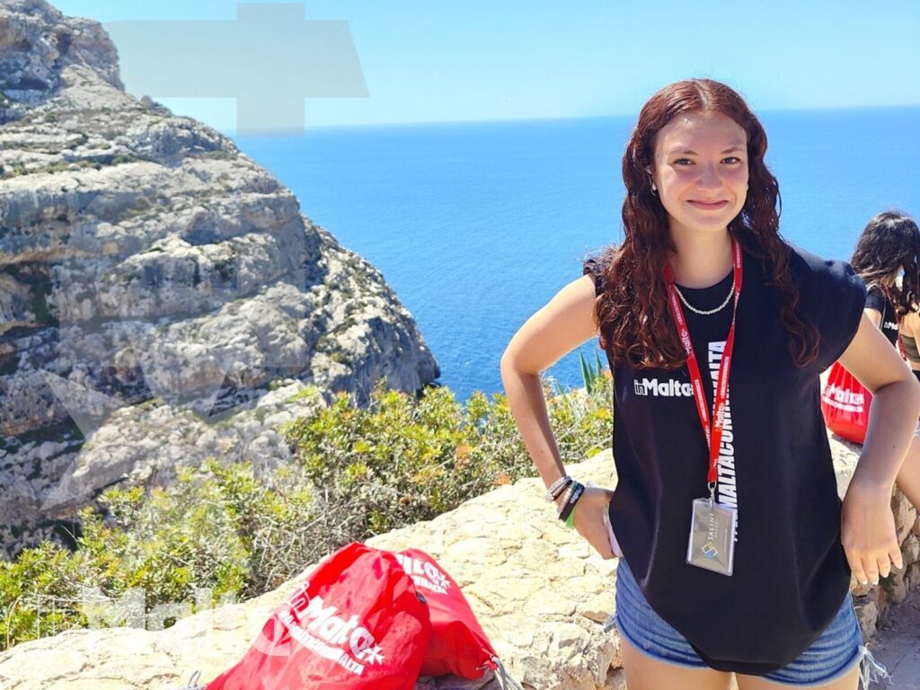 Studentessa durante l'escursione alla Blue Grotto di Malta Studentessa con maglietta del programma e tesserino identificativo posa davanti alla scogliera e al mare azzurro della costa maltese.
