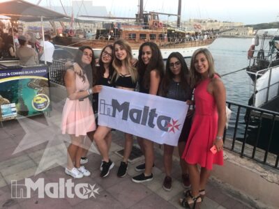 Sei studentesse sorridenti mostrano uno striscione inMalta sul lungomare, con barche e porto turistico sullo sfondo.