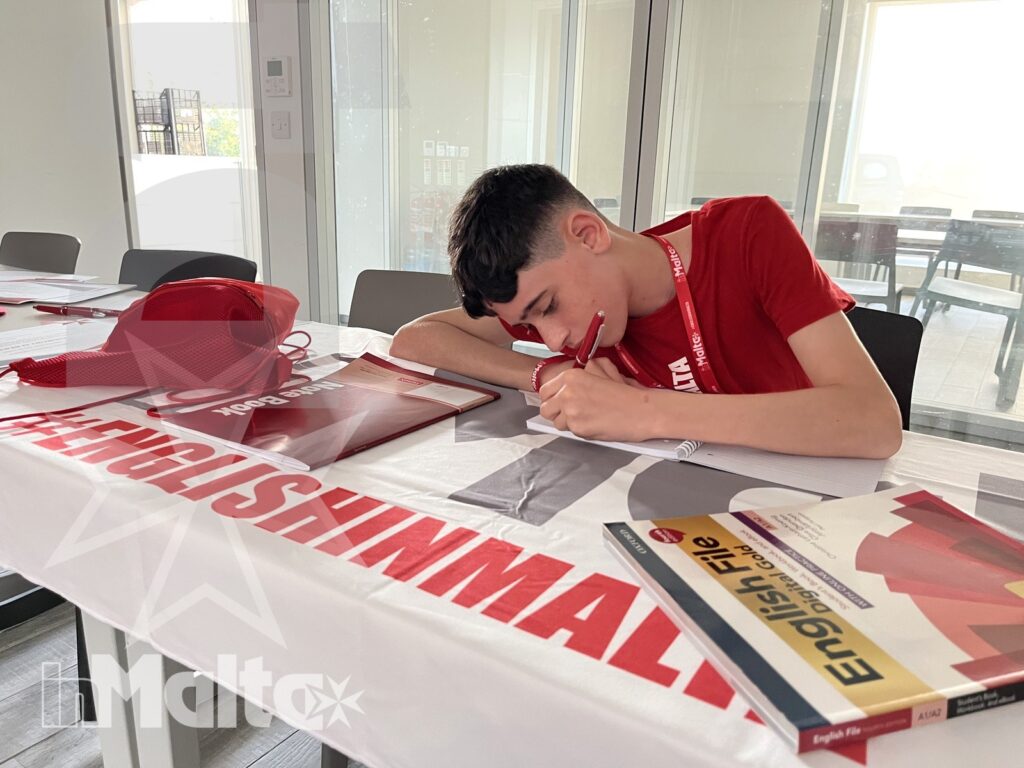 Giovane studente compila un modulo con penna, libro di inglese e cartellina rossa durante un'attività formativa inMalta.