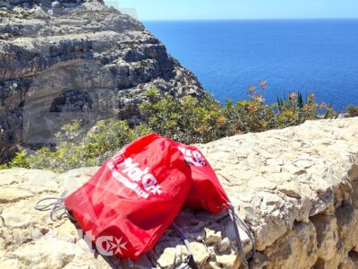 Scogliera rocciosa e mare aperto con vegetazione mediterranea e uno zaino rosso, atmosfera di viaggio sulla costa maltese.