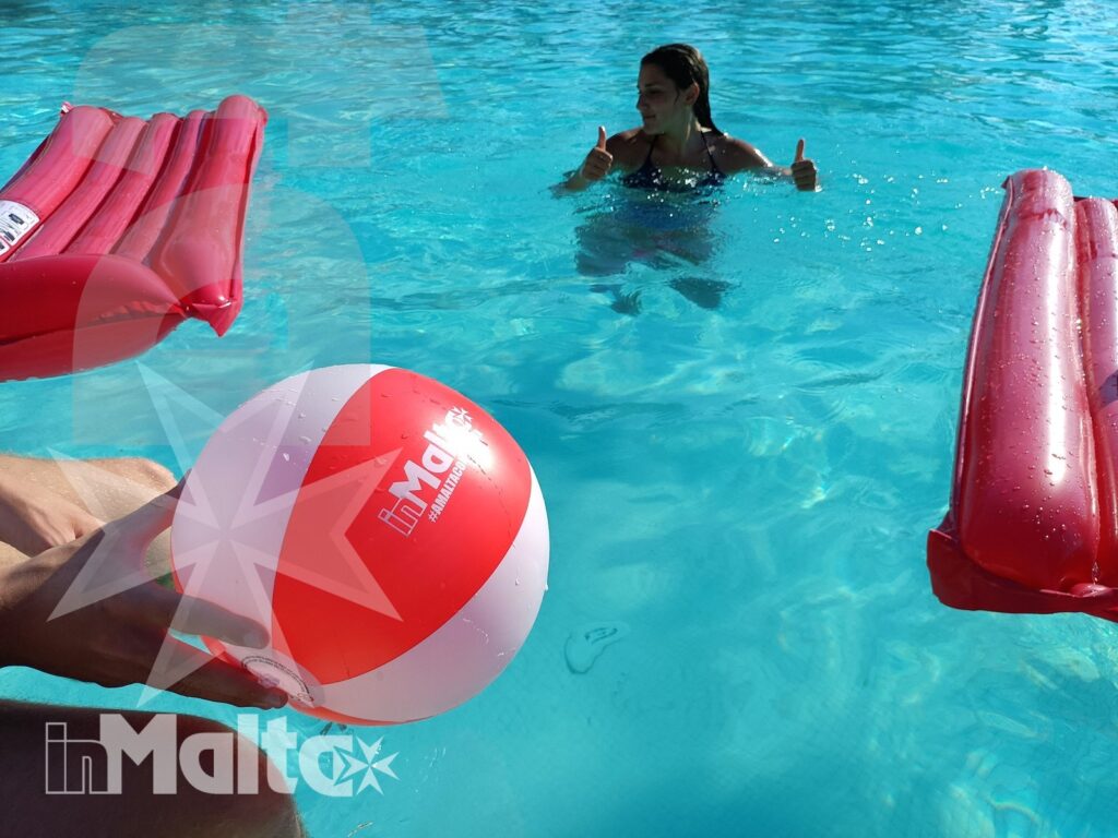 Uno studente in acqua fa un gesto positivo accanto a una palla da spiaggia e materassini rossi durante il soggiorno inMalta.