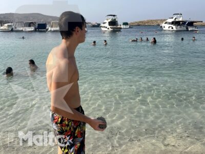 Uno studente passeggia sul bagnasciuga di Santa Maria Beach, con bagnanti e barche ancorate nella baia maltese.
