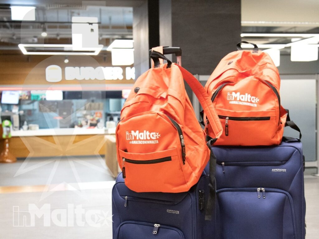 Zaini personalizzati e valigie per il viaggio a Malta Due zaini arancioni personalizzati sono appoggiati su valigie blu nell'area aeroportuale, pronti per un soggiorno linguistico a Malta.