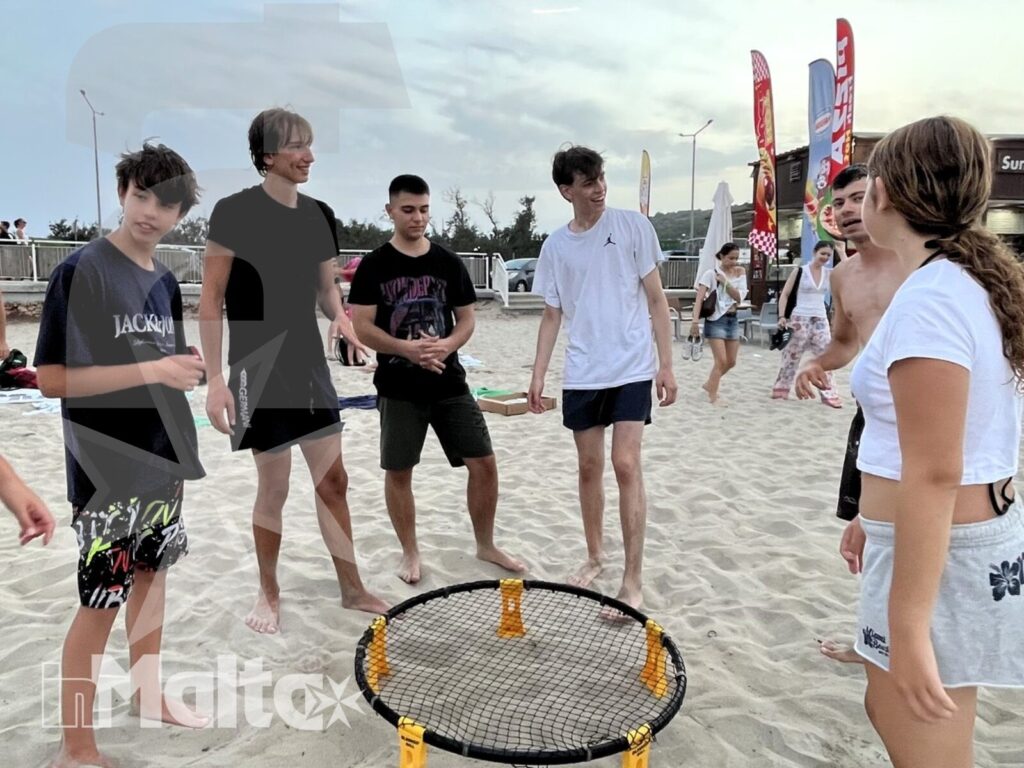 Studenti riuniti attorno a una rete da roundnet sulla sabbia, impegnati in un gioco di gruppo tra bandiere colorate.