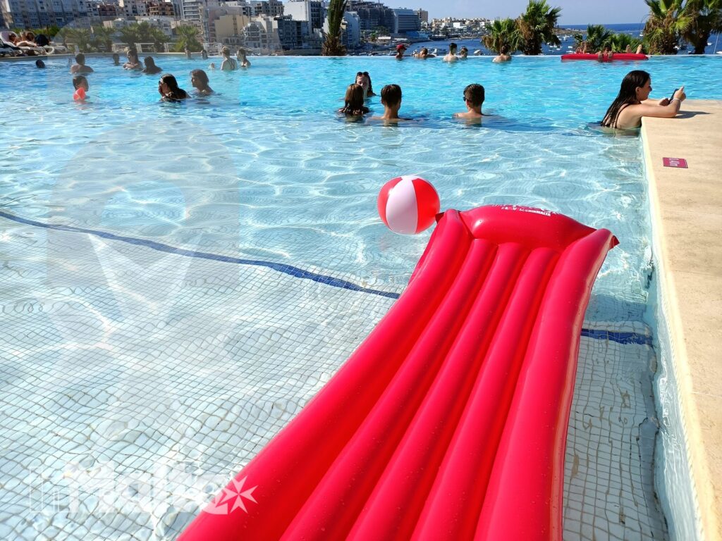 Materassino rosso in primo piano, bagnanti in piscina e panorama urbano costiero raccontano una giornata di relax degli studenti a Malta.