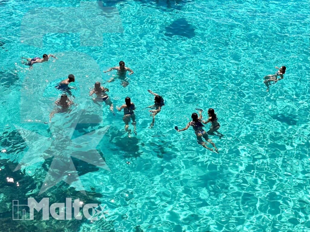 Gruppo di studenti nuota e gioca in una piscina dall'acqua turchese, durante un'attività ricreativa del soggiorno studio.